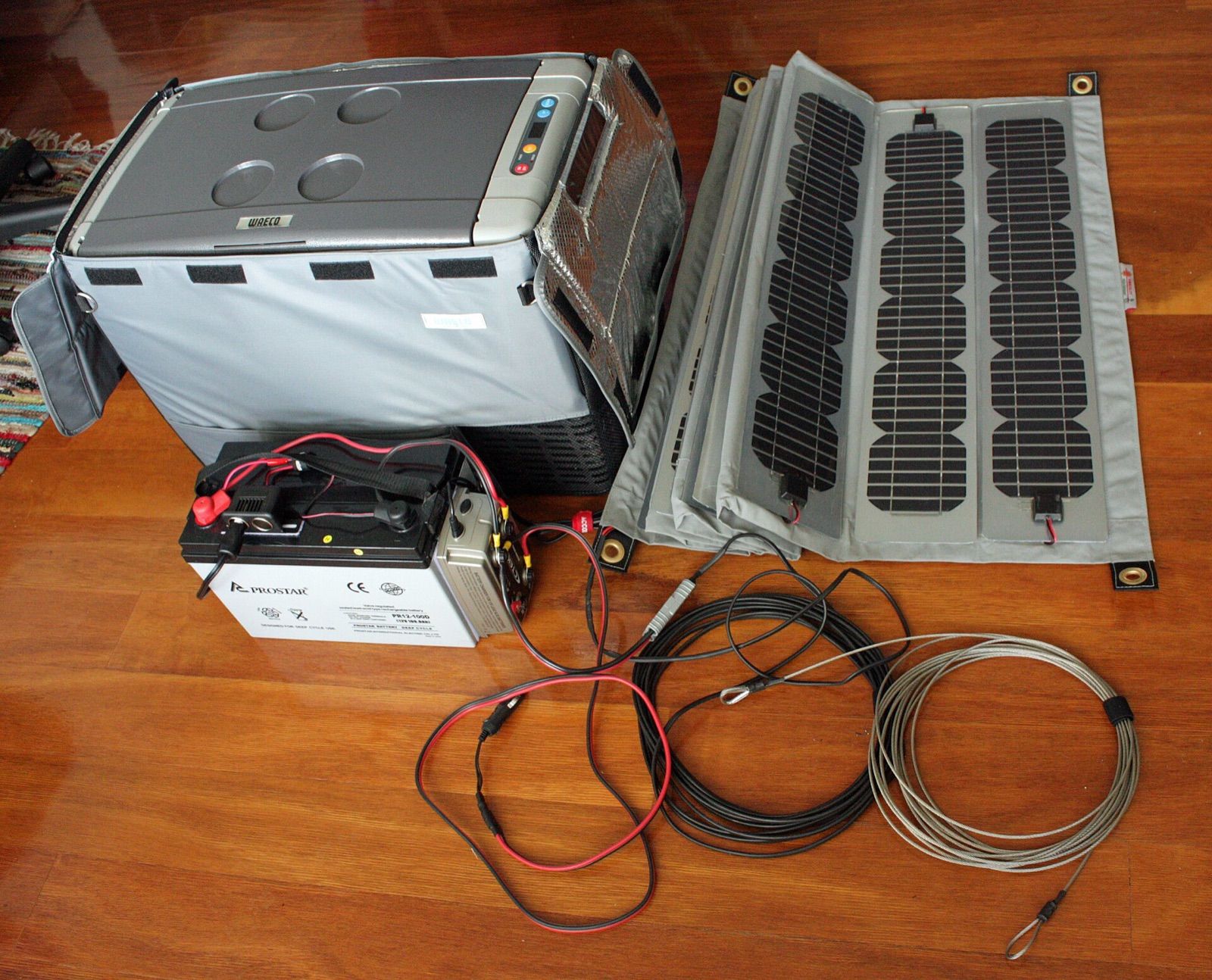 A portable 12 V fridge in an insulated cover beside a leisure battery and a folding solar panel