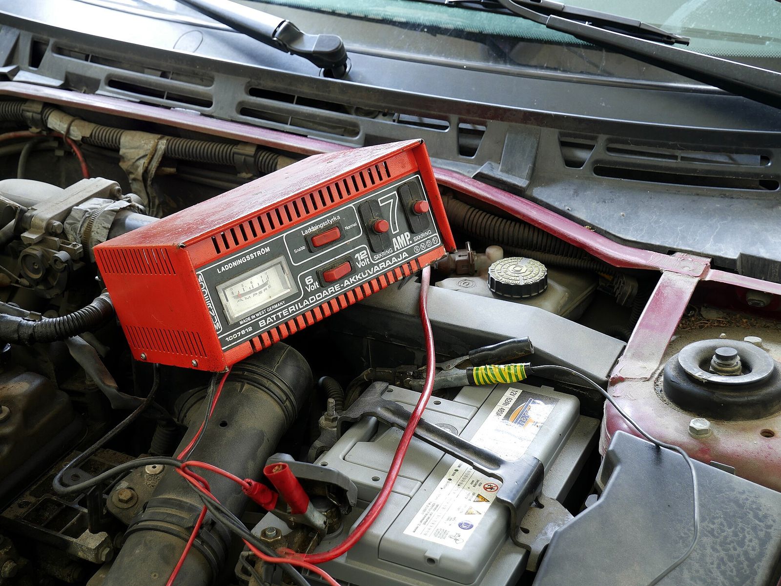 A car battery in an engine bay with the clamps of an old charger connected to its terminals