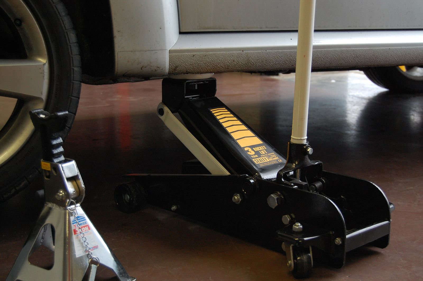 A low-profile floor jack positioned under a car's sill next to a front wheel, with a jack stand on the garage floor