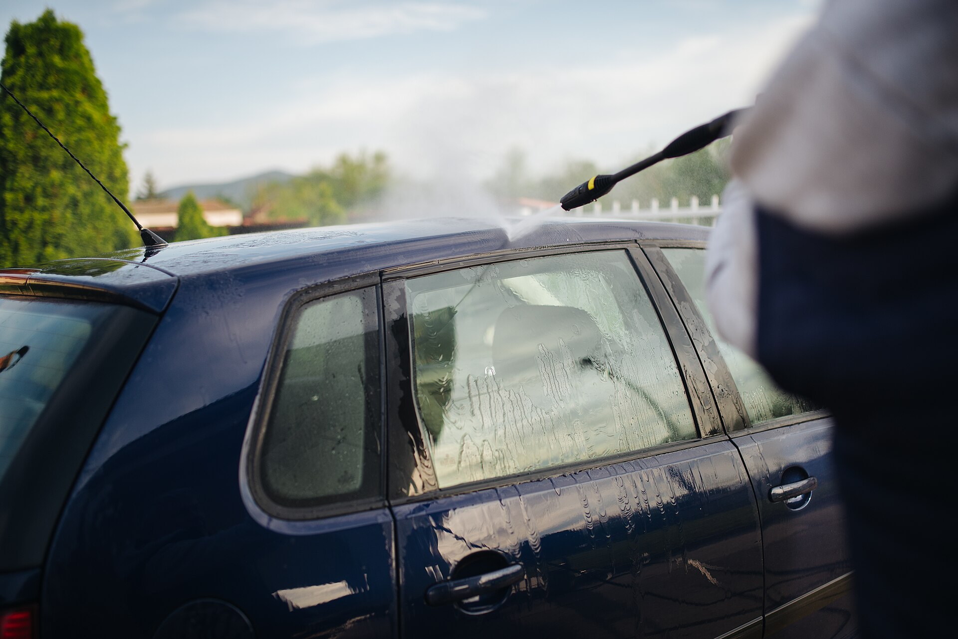 A person rinsing a blue hatchback with a pressure washer on a home driveway
