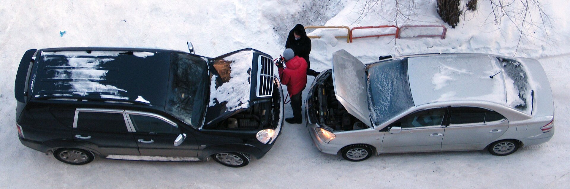 Two cars parked nose to nose in the snow with bonnets open while one jump-starts the other