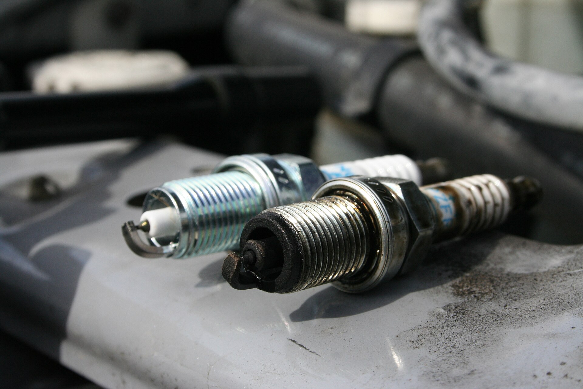 A new spark plug beside a used one with a darkened electrode, resting on an engine
