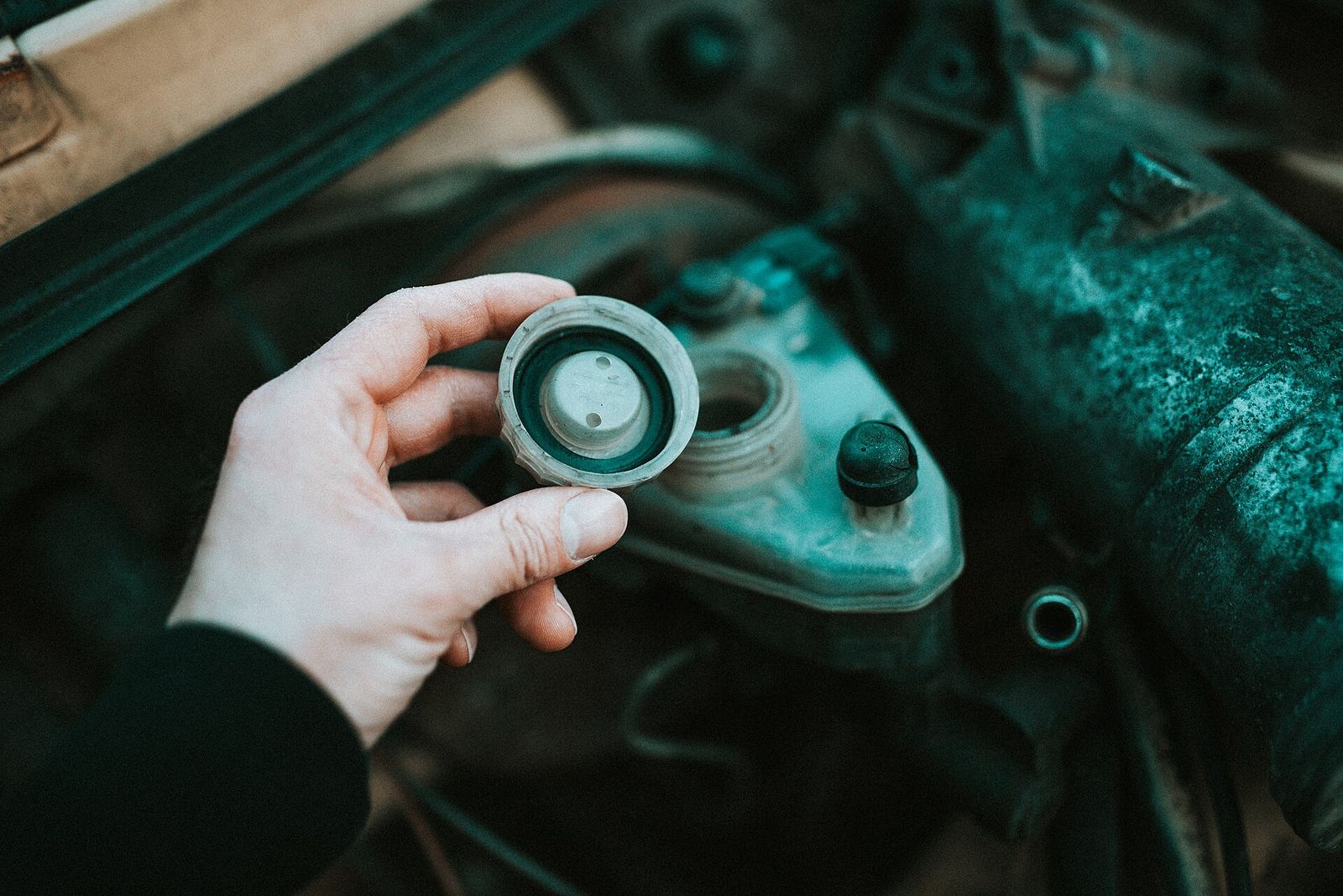 A hand holding a reservoir cap over an open fluid reservoir in a car's engine bay