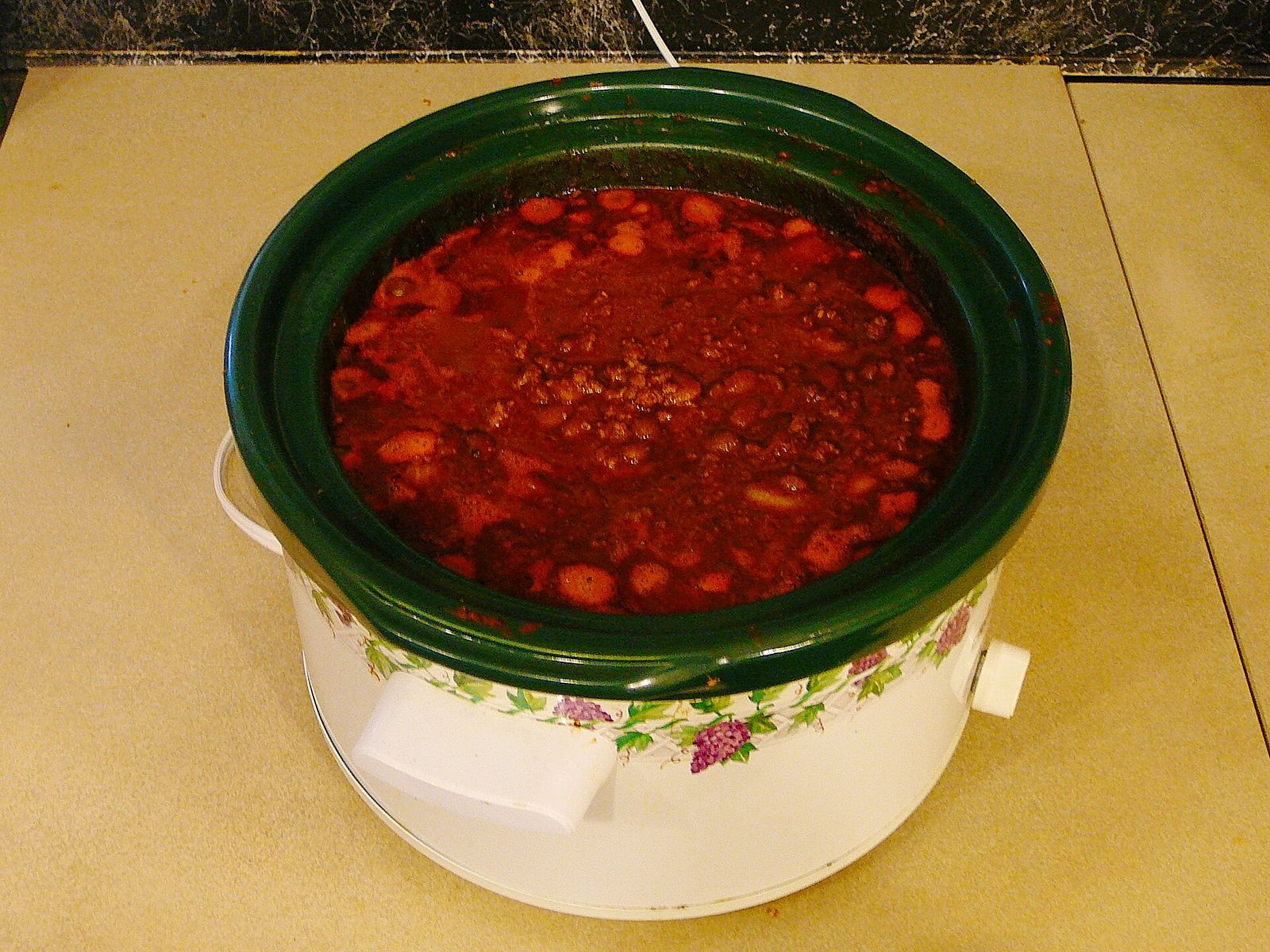 A round slow cooker with a dark green stoneware pot filled with chili on a kitchen counter