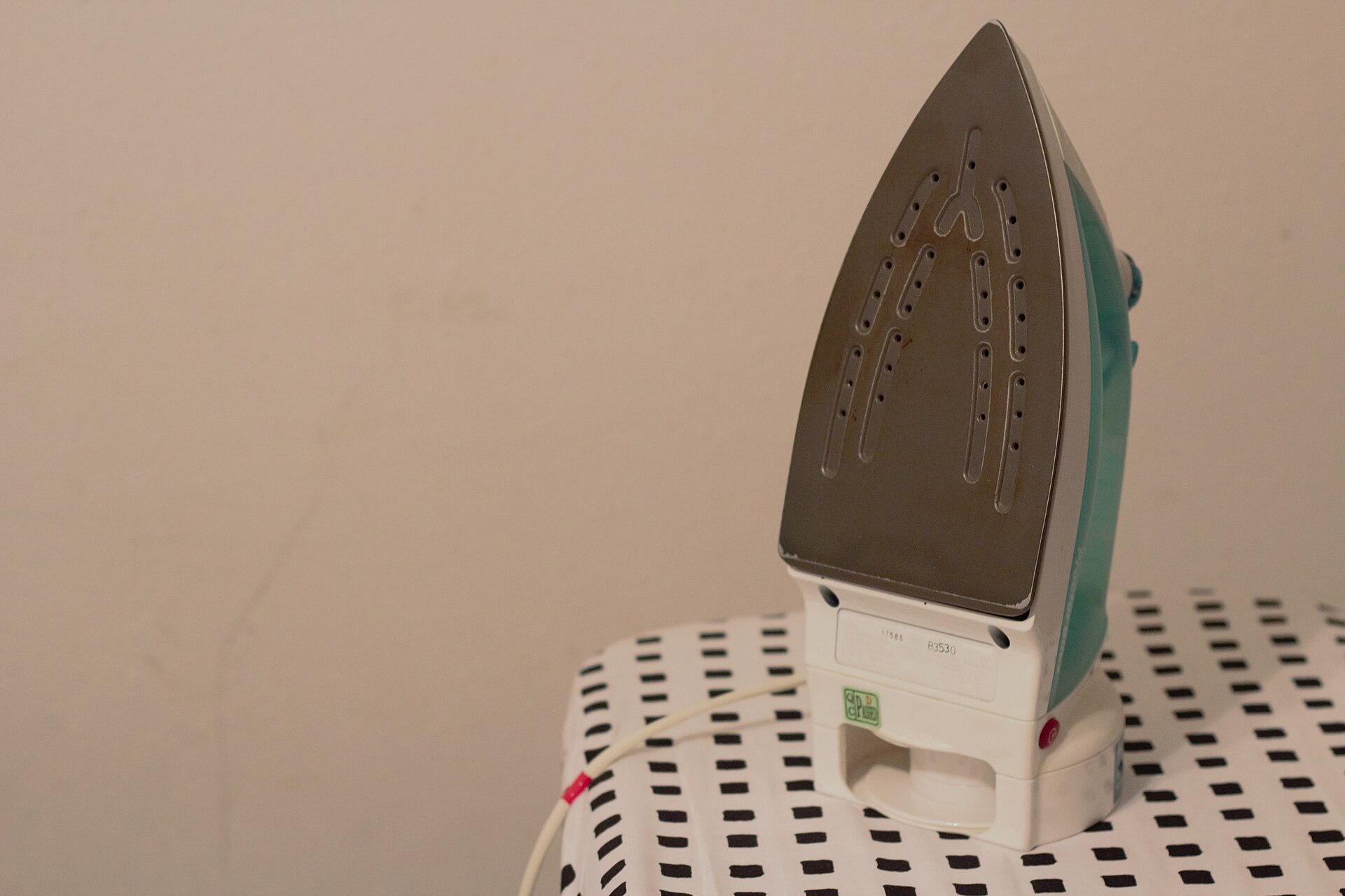 A steam iron standing on its heel on a patterned ironing board, showing its vented soleplate
