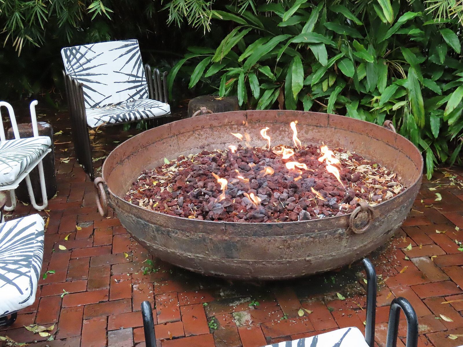 Gas fire bowl filled with lava rock and small flames on a brick patio, with garden chairs around it