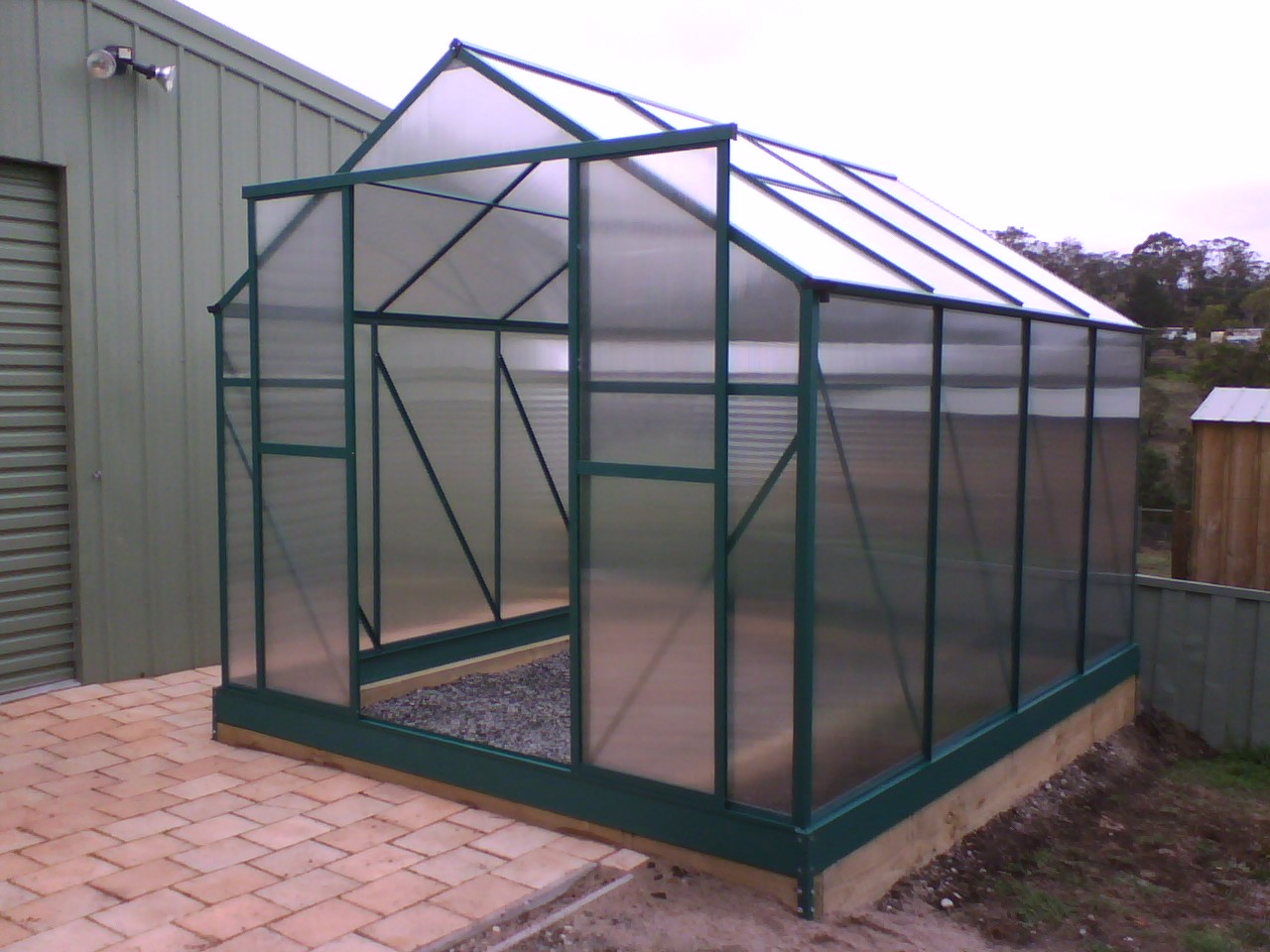 A small green-framed polycarbonate greenhouse on a timber base beside a paved path
