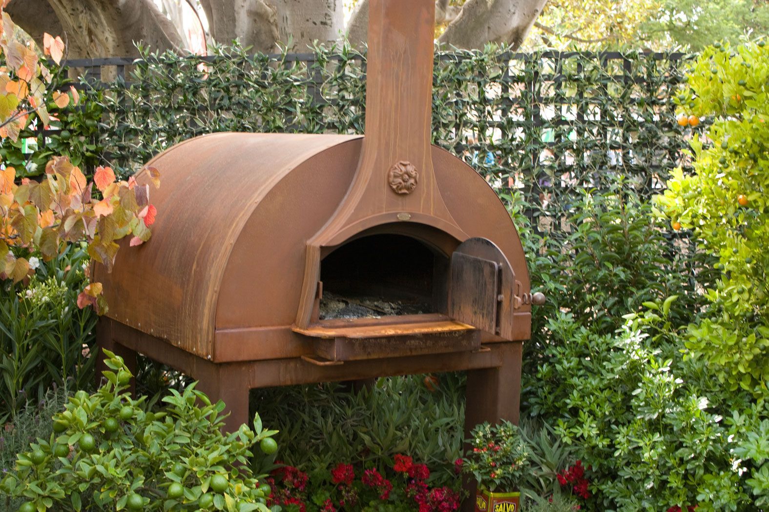 A dome-shaped metal wood-fired pizza oven surrounded by plants in a garden