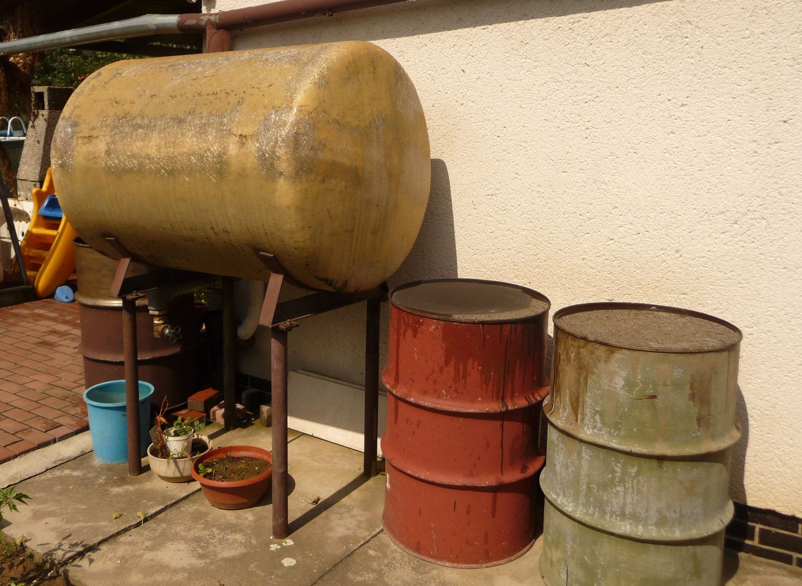 Rainwater tank on a stand below a gutter, with storage drums beside a house wall