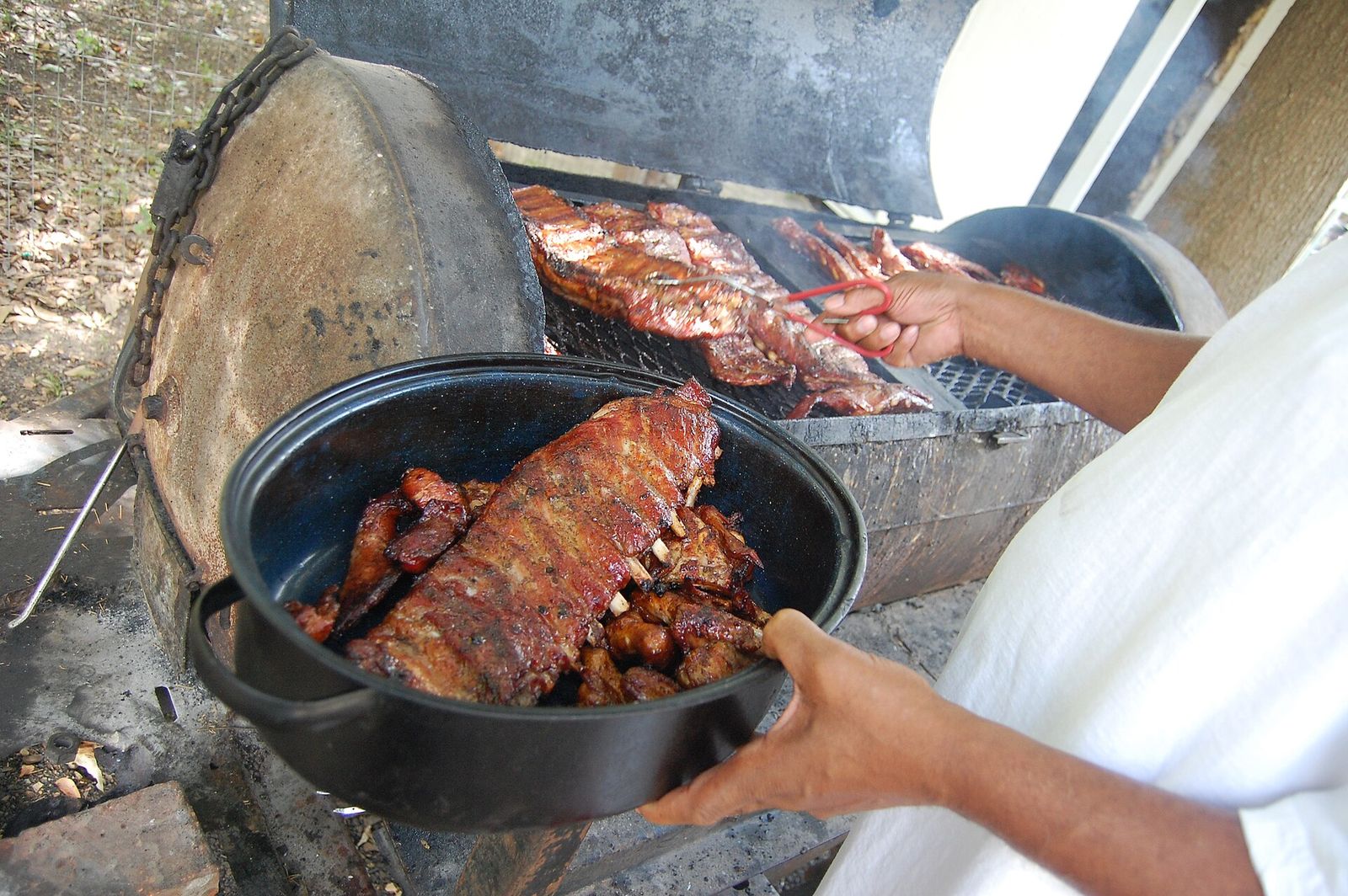 Racks of pork ribs being lifted from a large steel barrel smoker into a roasting pan