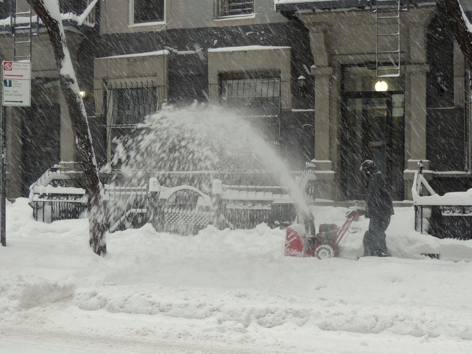 A person clearing a snowy city sidewalk with a snow blower during a heavy snowstorm