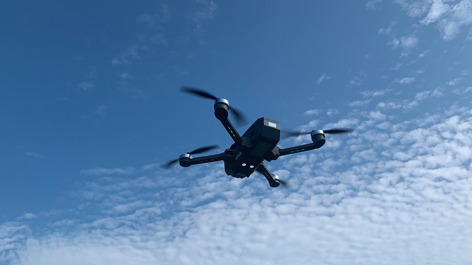 A small folding camera drone with four propellers flying against a blue sky with scattered clouds