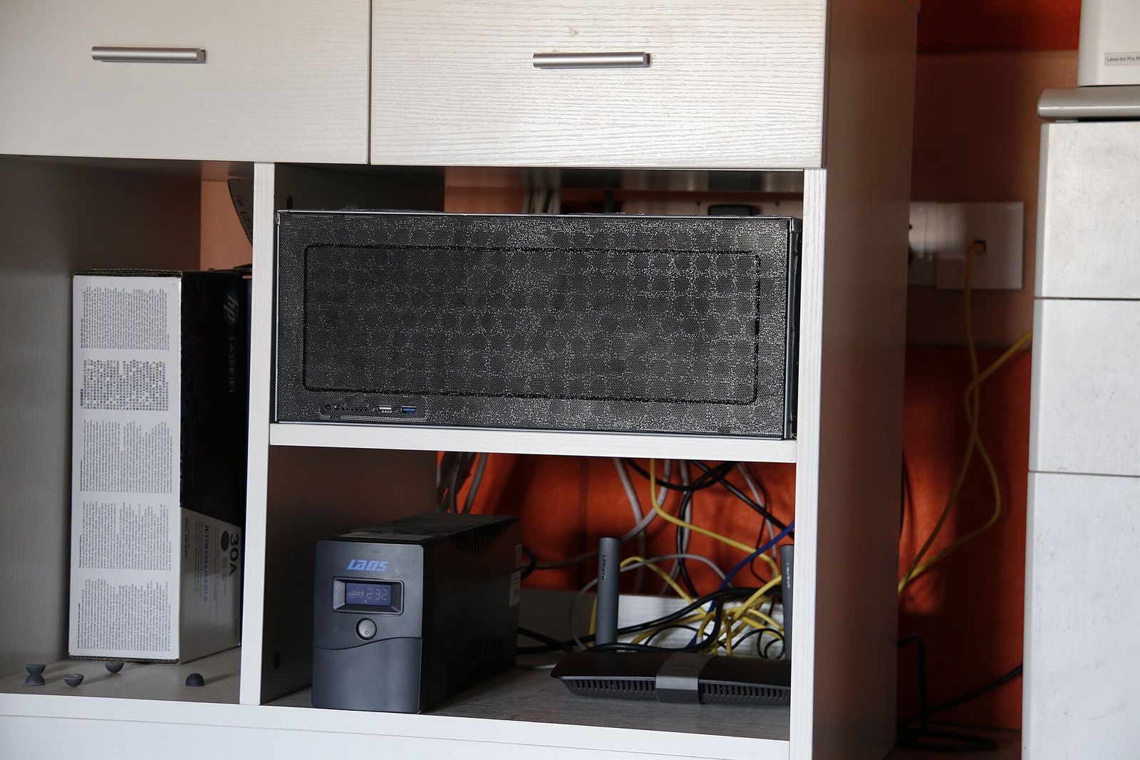 A compact home server on a shelf above a small uninterruptible power supply and a Wi-Fi router