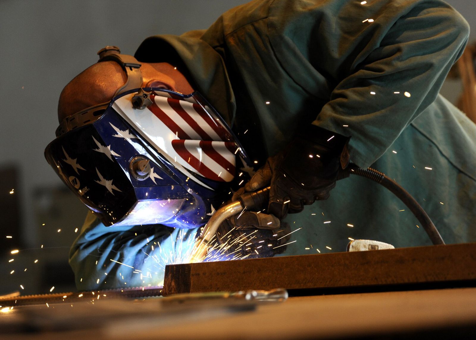 A welder in an auto-darkening helmet and leather gloves MIG welding a steel bar, with sparks flying