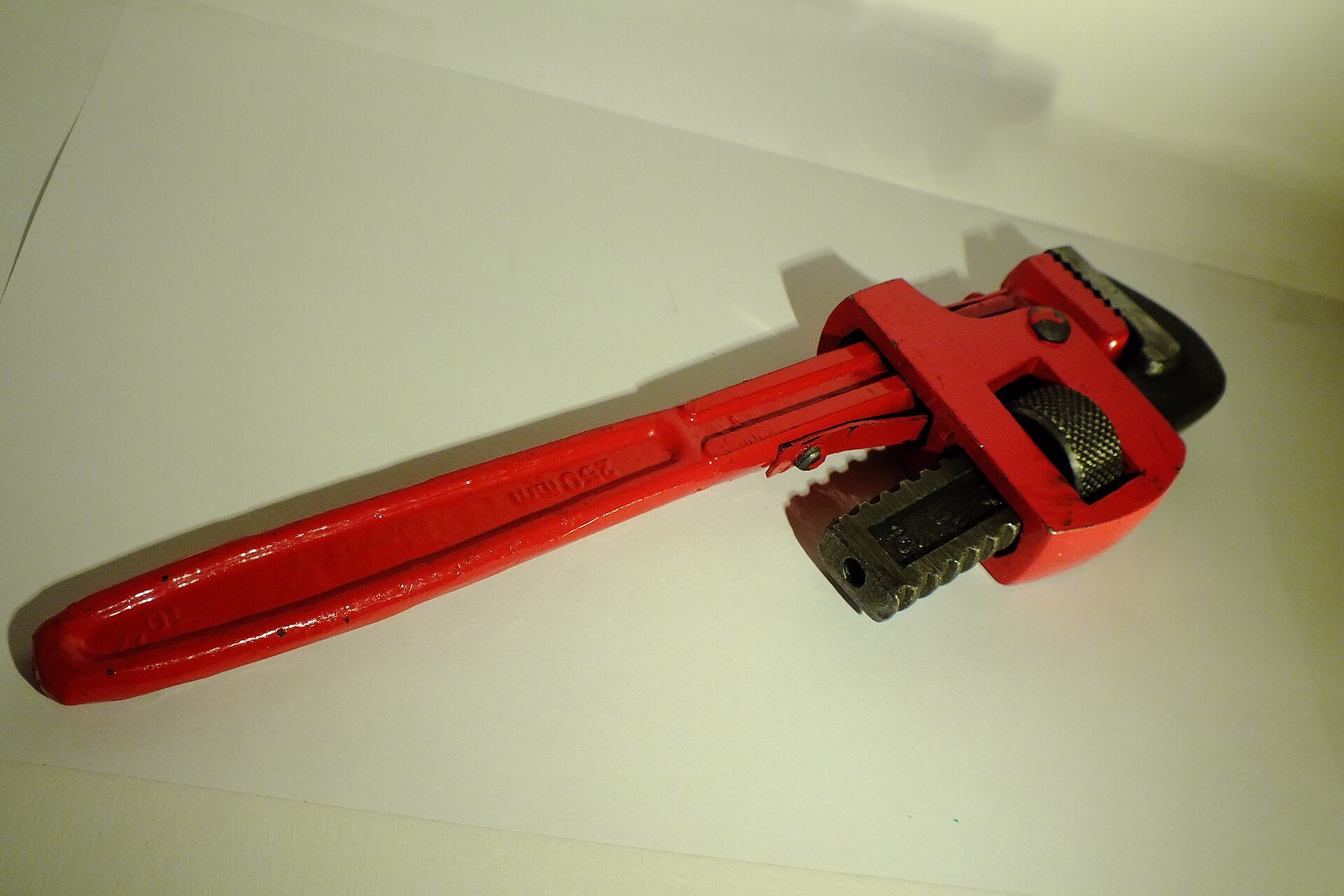 A red adjustable pipe wrench with serrated steel jaws lying on a sheet of white paper