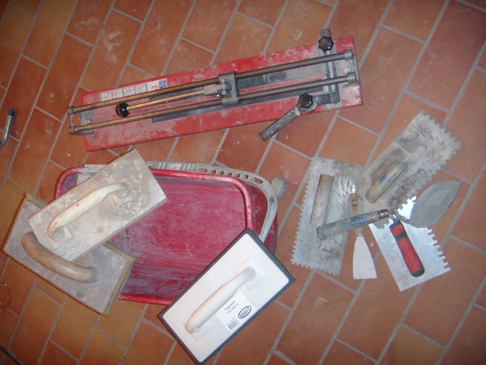 A manual tile cutter, notched trowels, a grout float and a mixing tub laid out on a tiled floor