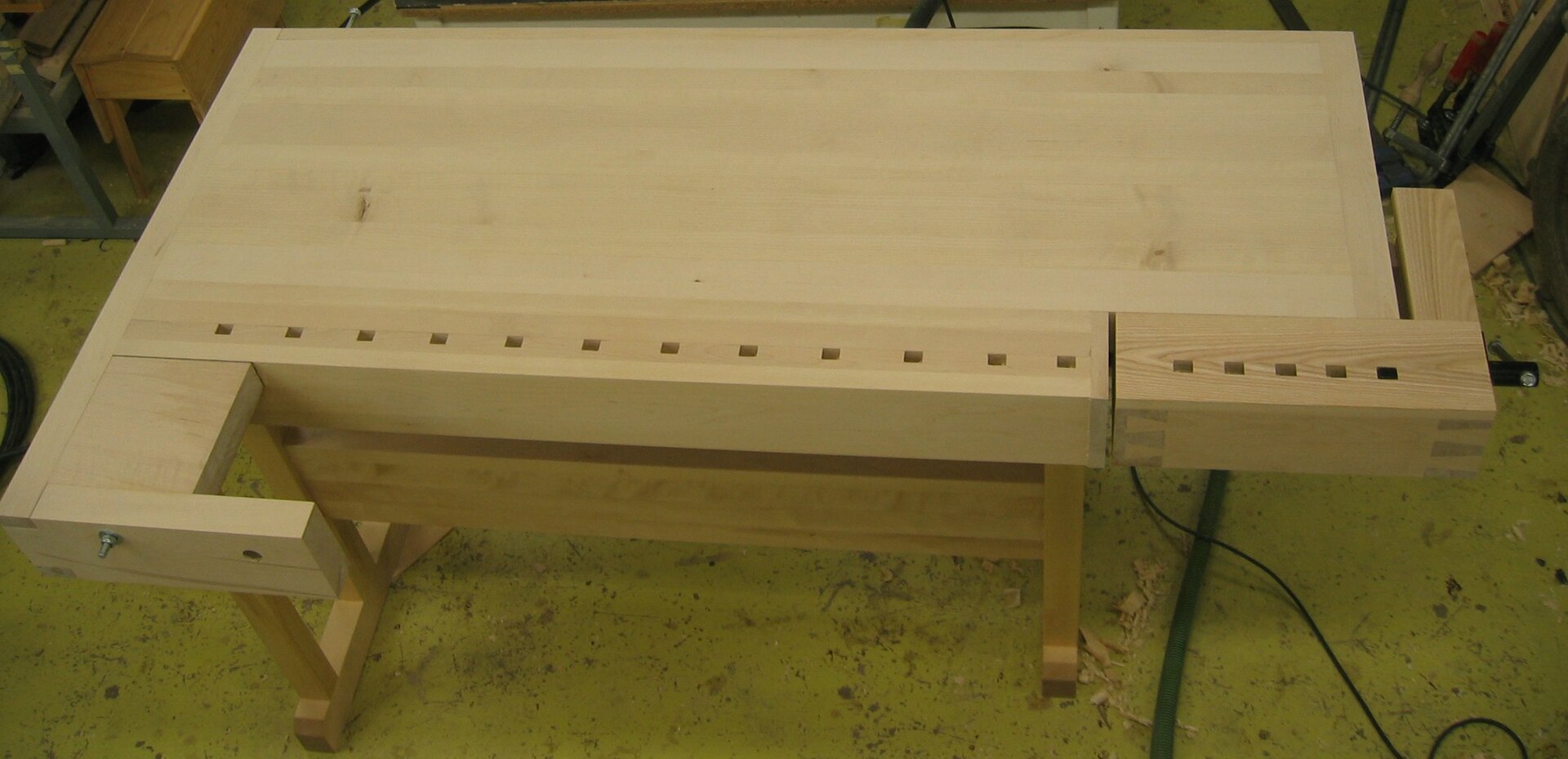 A solid wooden woodworking bench with a row of dog holes and a wooden vise at one end
