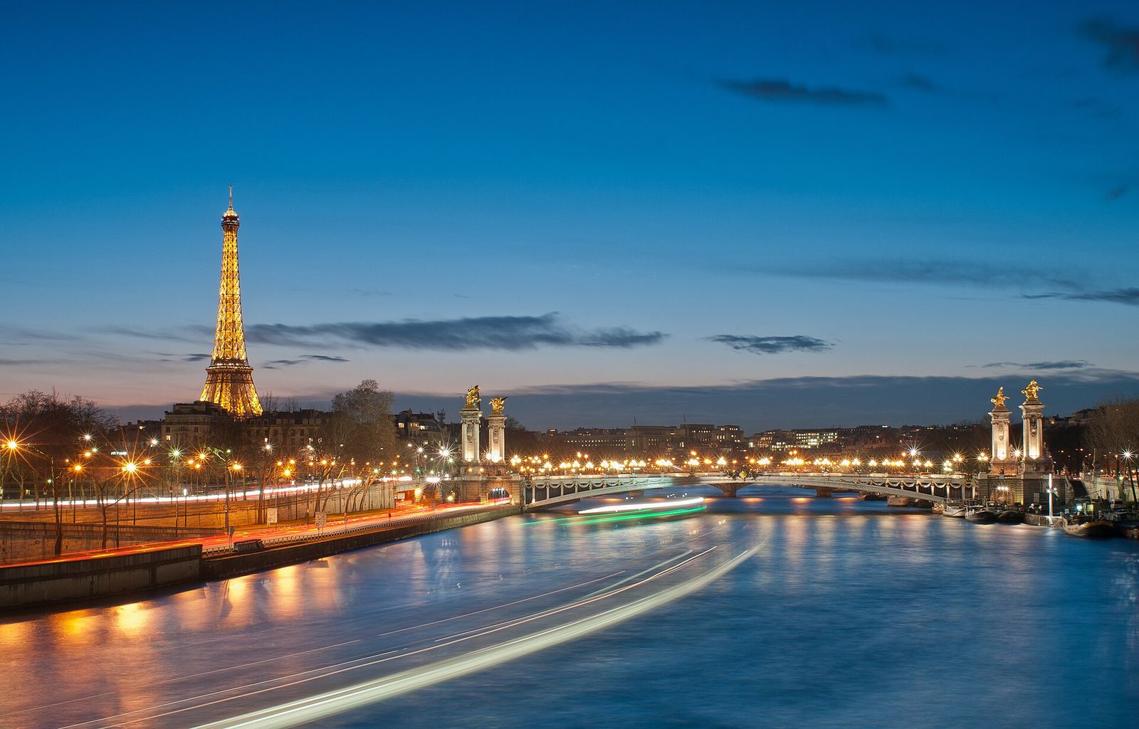 The Eiffel Tower lit up at dusk beyond the Pont Alexandre III and the Seine